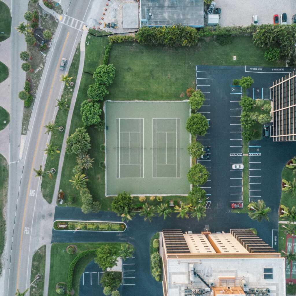 Overhead drone shot of a tennis court surrounded by greenery and parking in a cityscape.