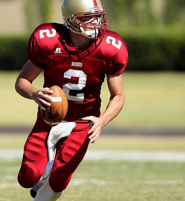 Quarterback in red jersey running with football on grassy field during daytime sporting event.